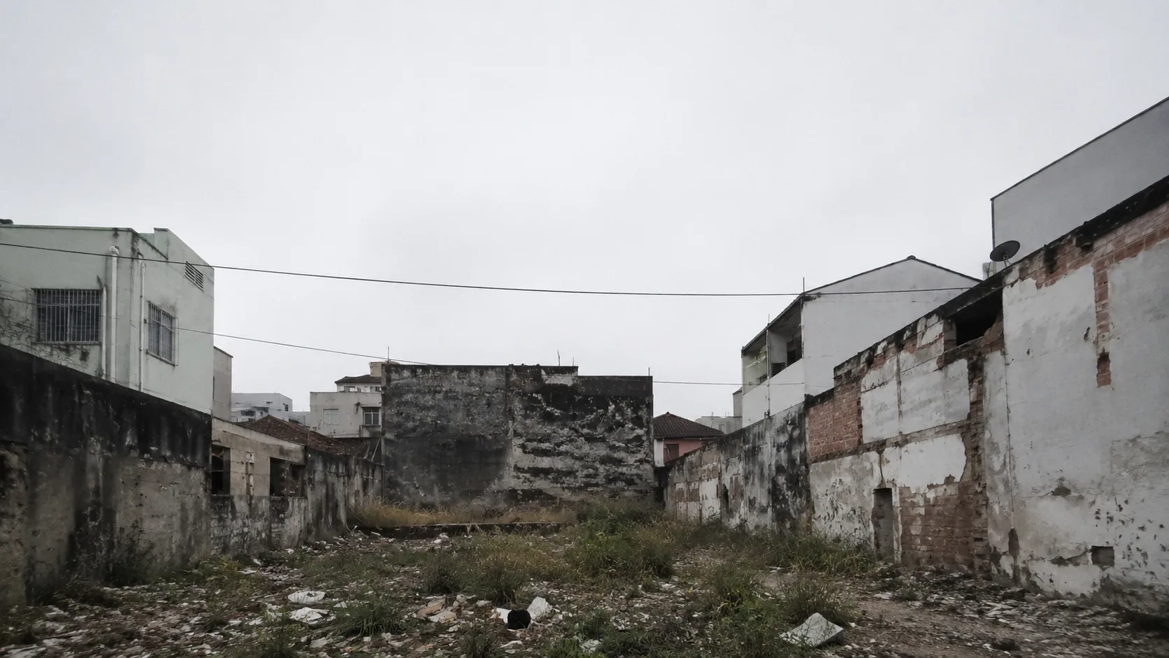 Terreno baldio abandonado entre empenas cegas, com mato e entulho sob céu encoberto, exemplo de ponto de vergonha em cidade do Sudeste do Brasil