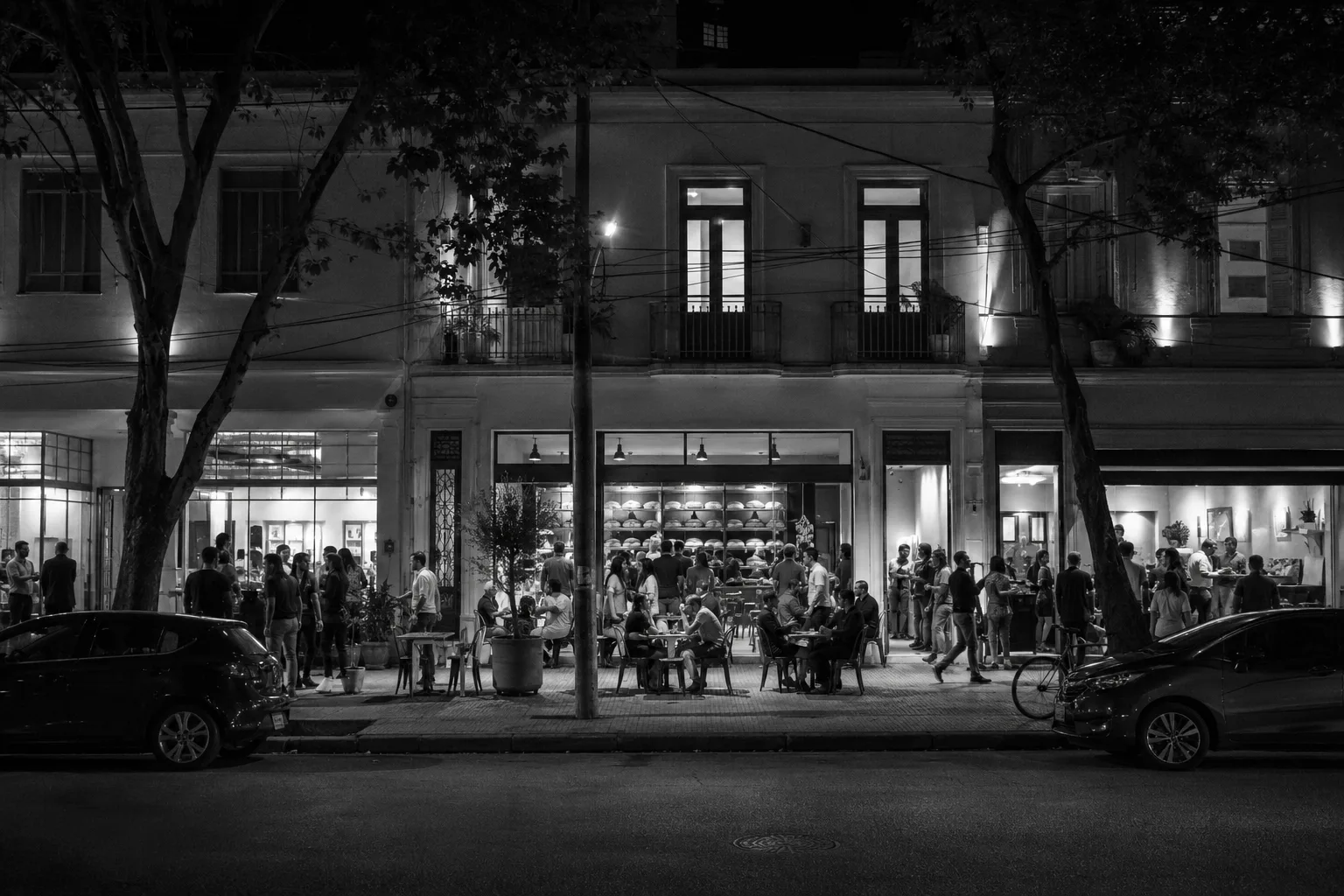 Rua de bairro à noite com restaurante de térreo cheio, mesas na calçada e apartamentos iluminados acima, presença contínua, cidade do Sudeste do Brasil